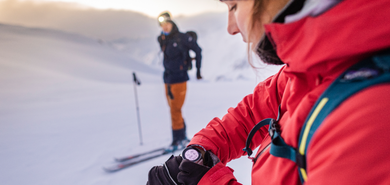 Two Female athletes in backcountry skiing, athlete red jacket reviewing data on Suunto watch